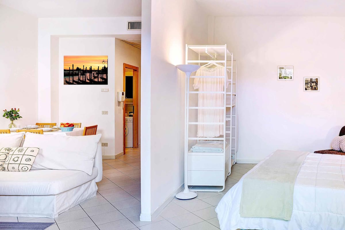 A hotel room with a white bed, a white wardrobe, a white desk, and a white chair. There is a window with a view of a building and a door leading to a bathroom.