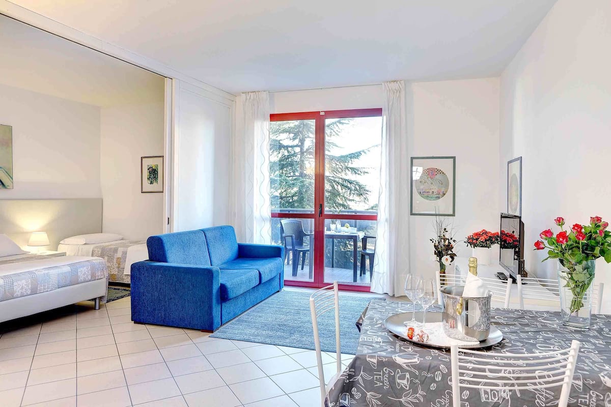 A modern room with a bed, a blue sofa, a dining table with chairs, and a view through a glass door to an outdoor area.