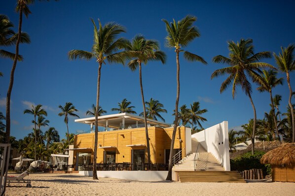 A modern beachside building with a wooden facade, outdoor seating, and palm trees.