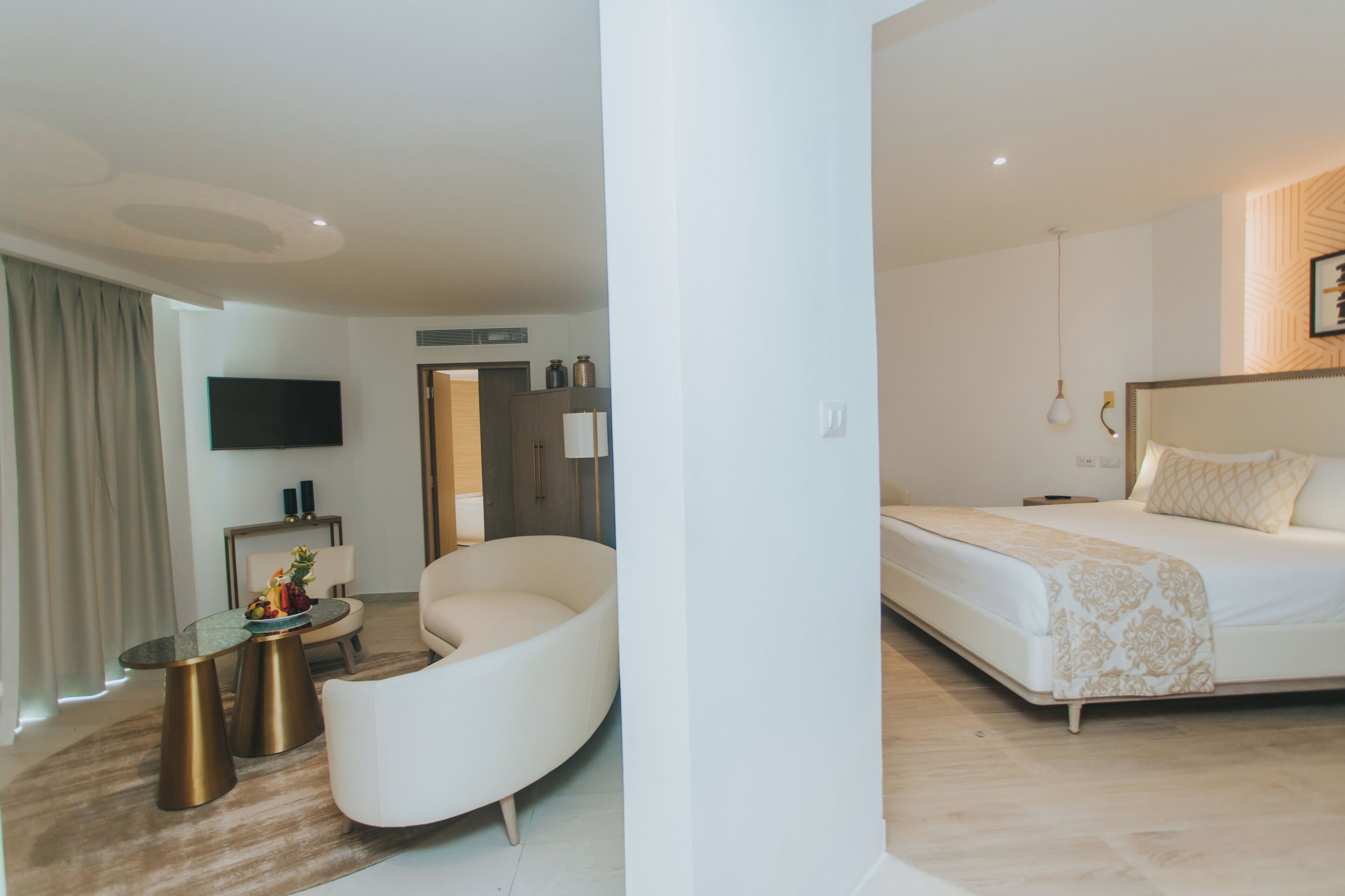 Una habitación de hotel moderna con un sofá blanco curvo, una mesa de centro de vidrio, un televisor de pantalla plana, una cama con colcha estampada y suelo de madera.