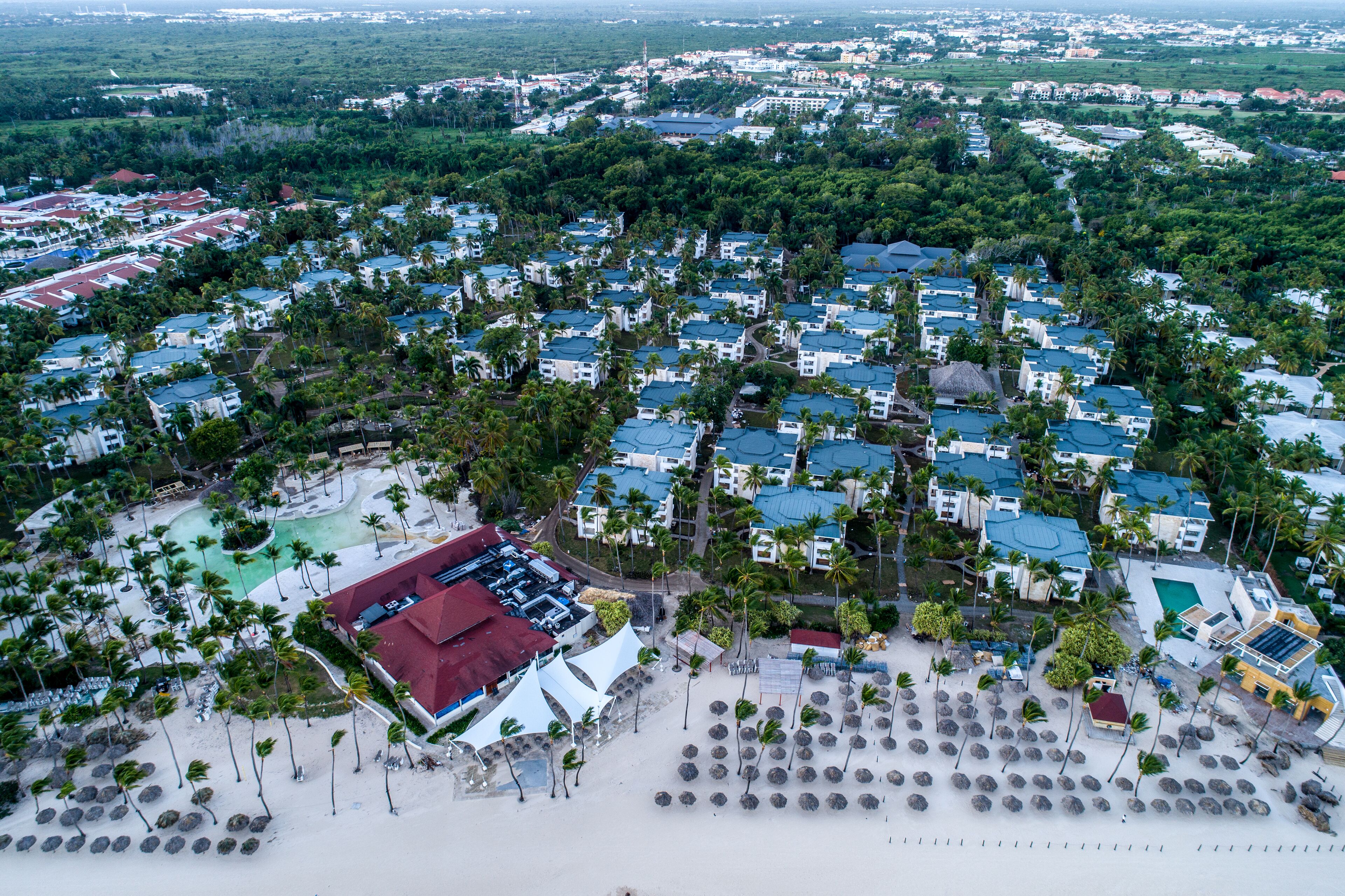 An aerial view of a resort with multiple buildings, a beach area with umbrellas, and a pool.