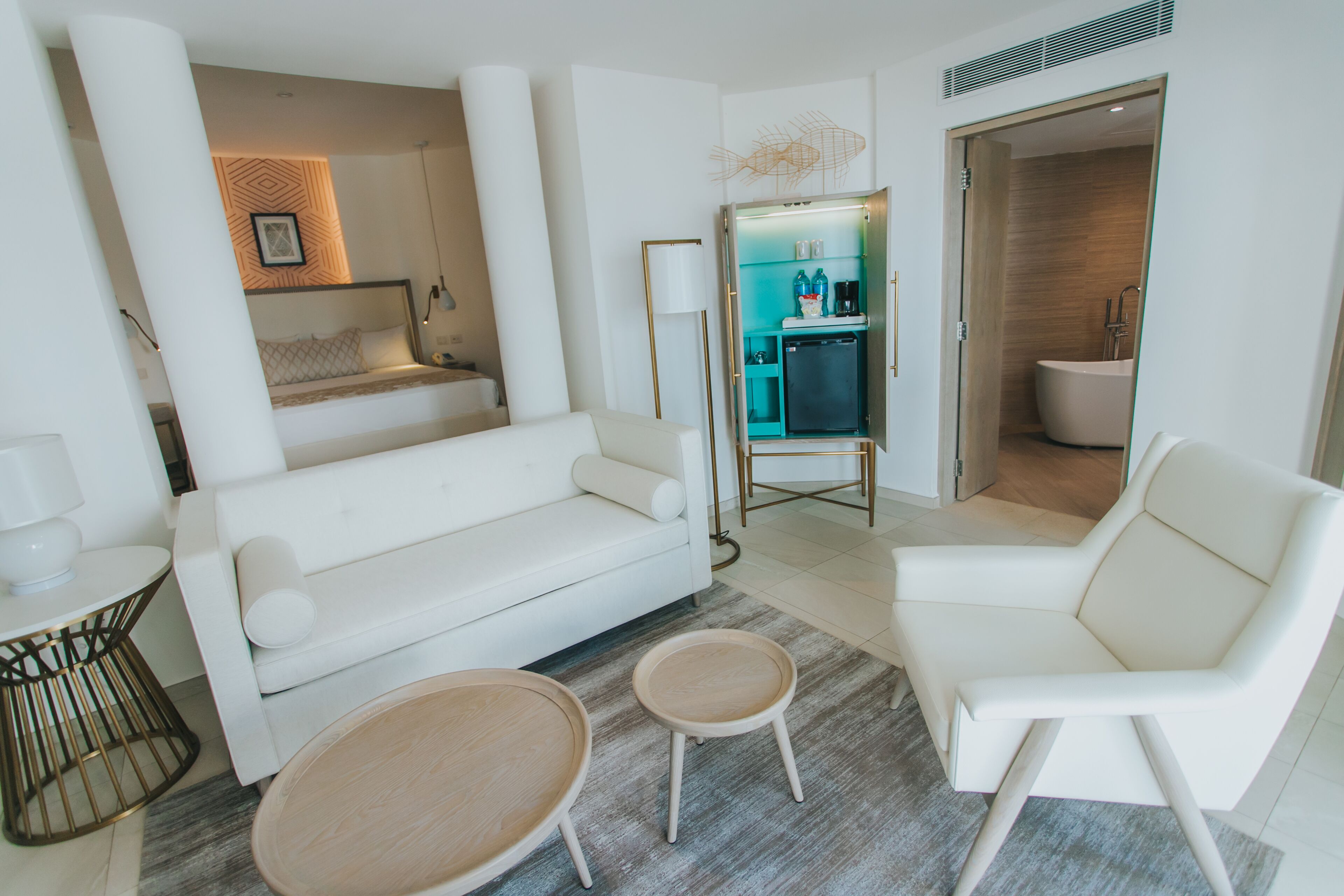 Un salon moderne avec un canapé blanc, deux petites tables en bois et un fauteuil blanc. Au fond, on aperçoit une petite cuisine équipée d’un réfrigérateur et d’un évier.