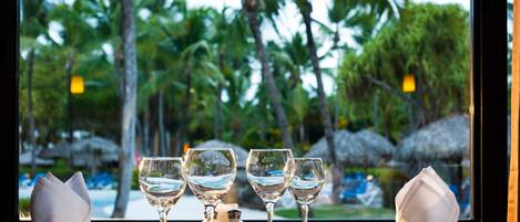 A table set with wine glasses and a salt and pepper shaker, viewed through a window overlooking palm trees and a beach.