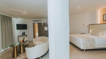 Una habitación de hotel moderna con un sofá blanco curvo, una mesa de centro de vidrio, un televisor de pantalla plana, una cama con colcha estampada y piso de madera.