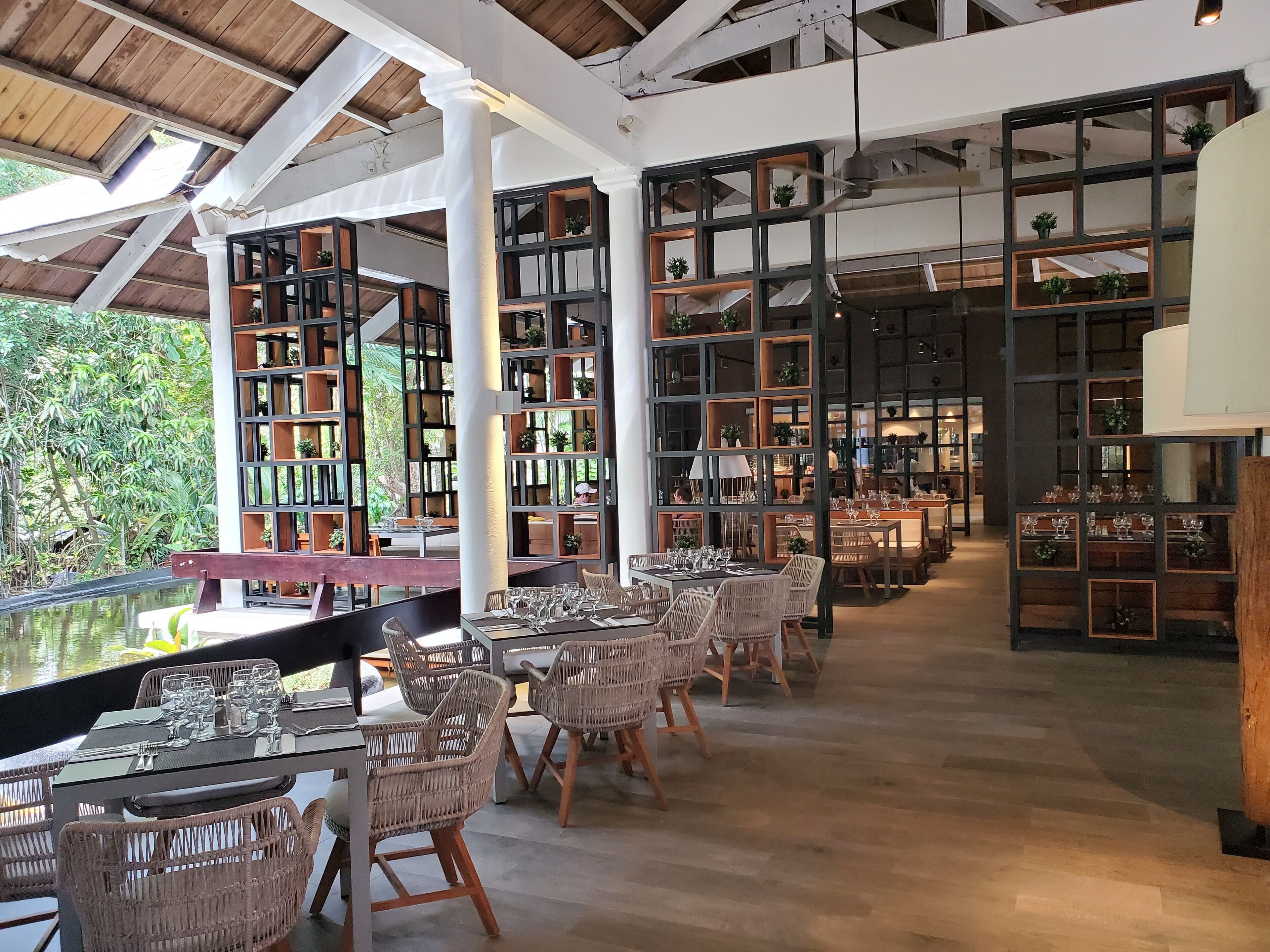 An indoor dining area with wicker chairs and tables, surrounded by tall bookshelves filled with books and plants.
