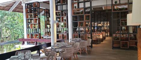 An indoor dining area with wicker chairs and tables, surrounded by tall bookshelves filled with books and plants.
