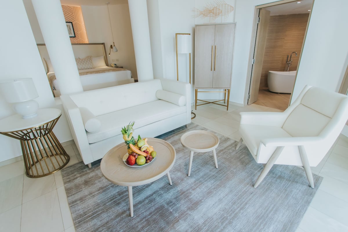 A modern living room with a white sofa, a round coffee table with fruit, a white armchair, and a small side table.