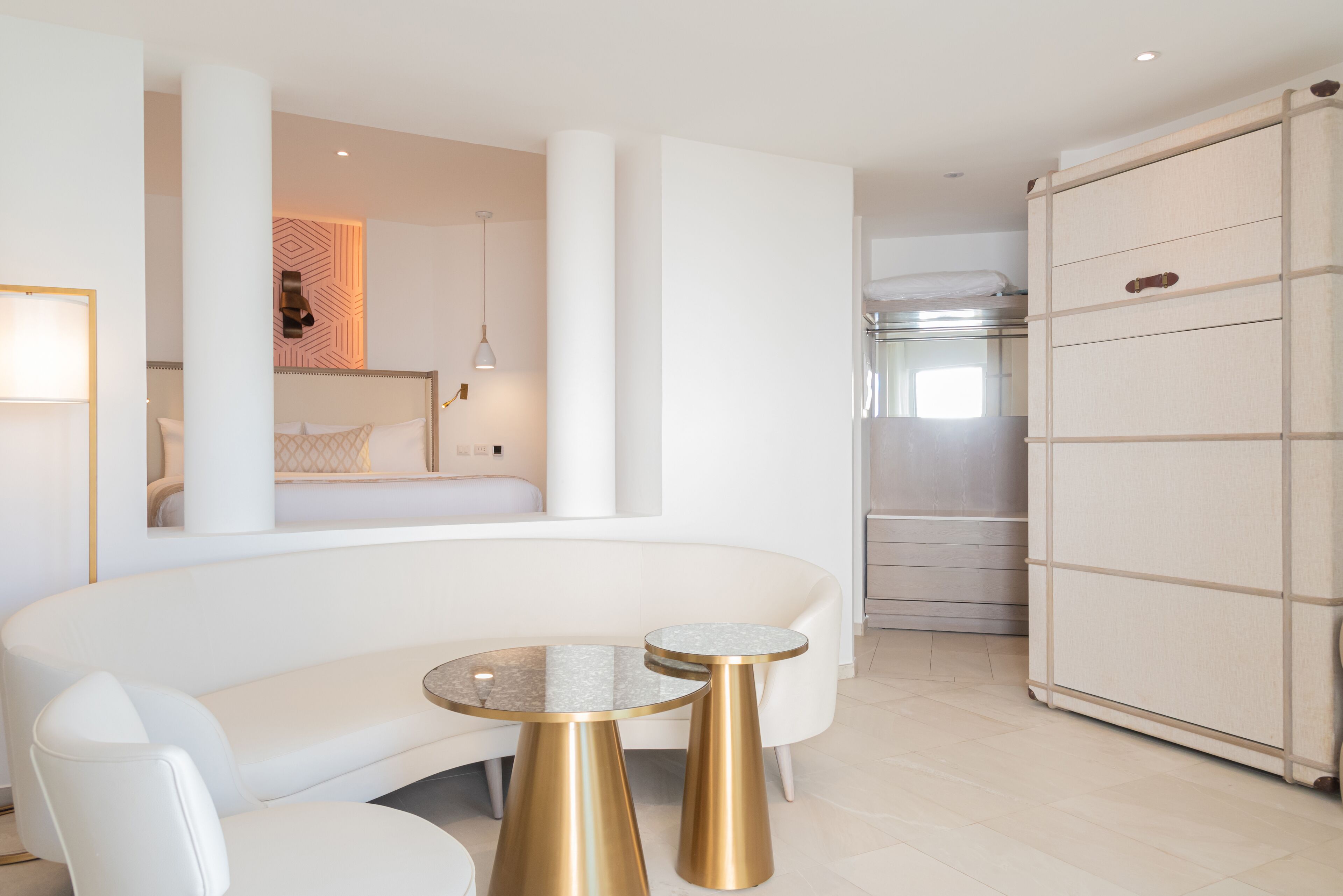 A modern hotel room with a white sofa, two gold-trimmed round tables, a large mirror, and a sliding wardrobe.