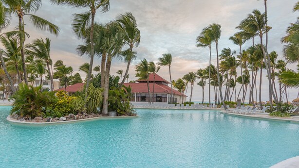 A pool area with palm trees and a building with a red roof. - Grand Bavaro Princess - All Inclusive (Punta Cana)