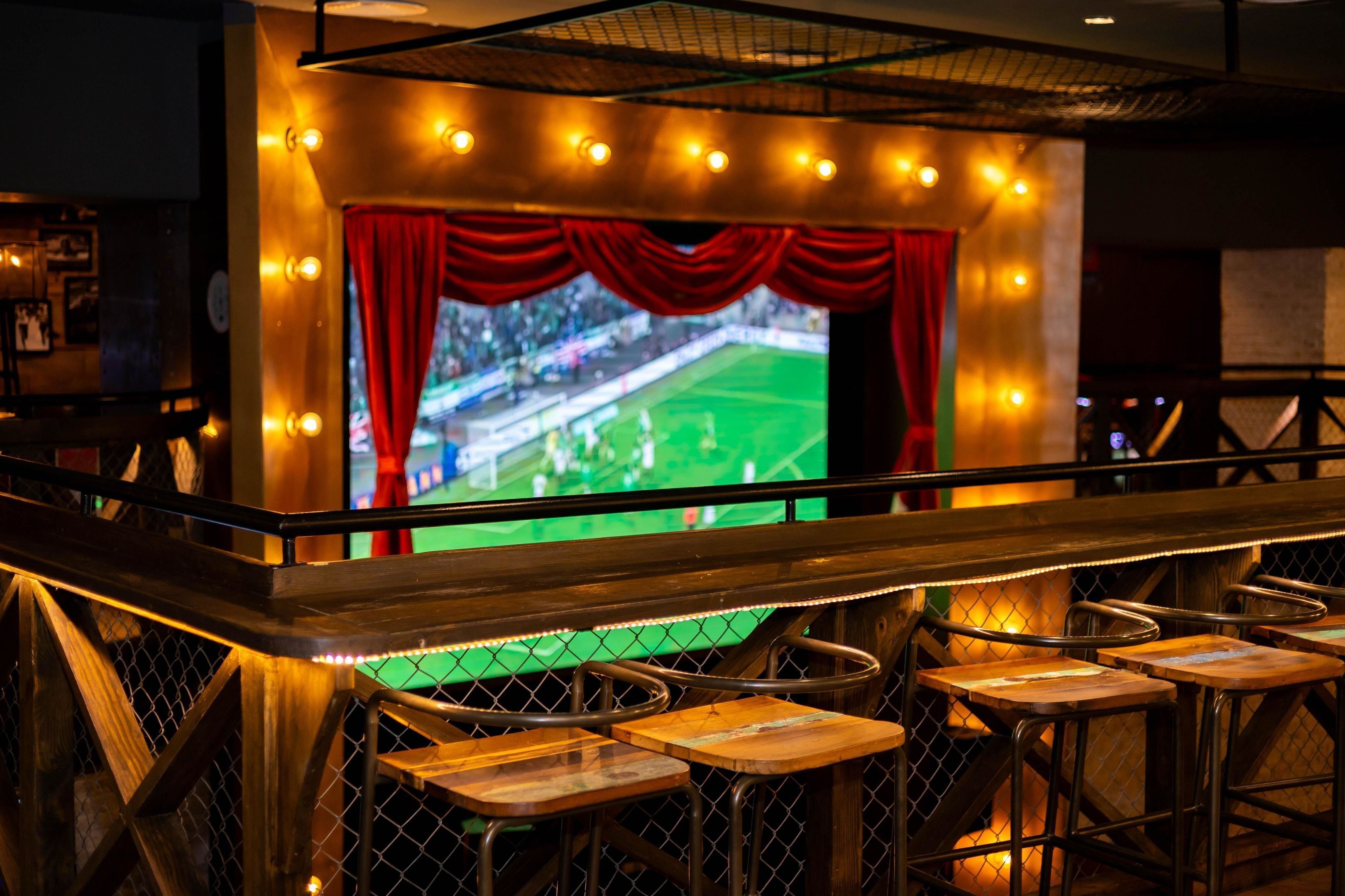 Un bar sportif doté d’un grand écran diffusant un match de football, de tabourets de bar en bois et d’une rambarde donnant sur l’espace réservé aux clients.