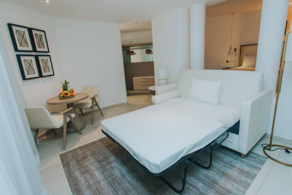 A modern hotel room with a bed, a small dining table with chairs, a sofa, and a view of the kitchen.