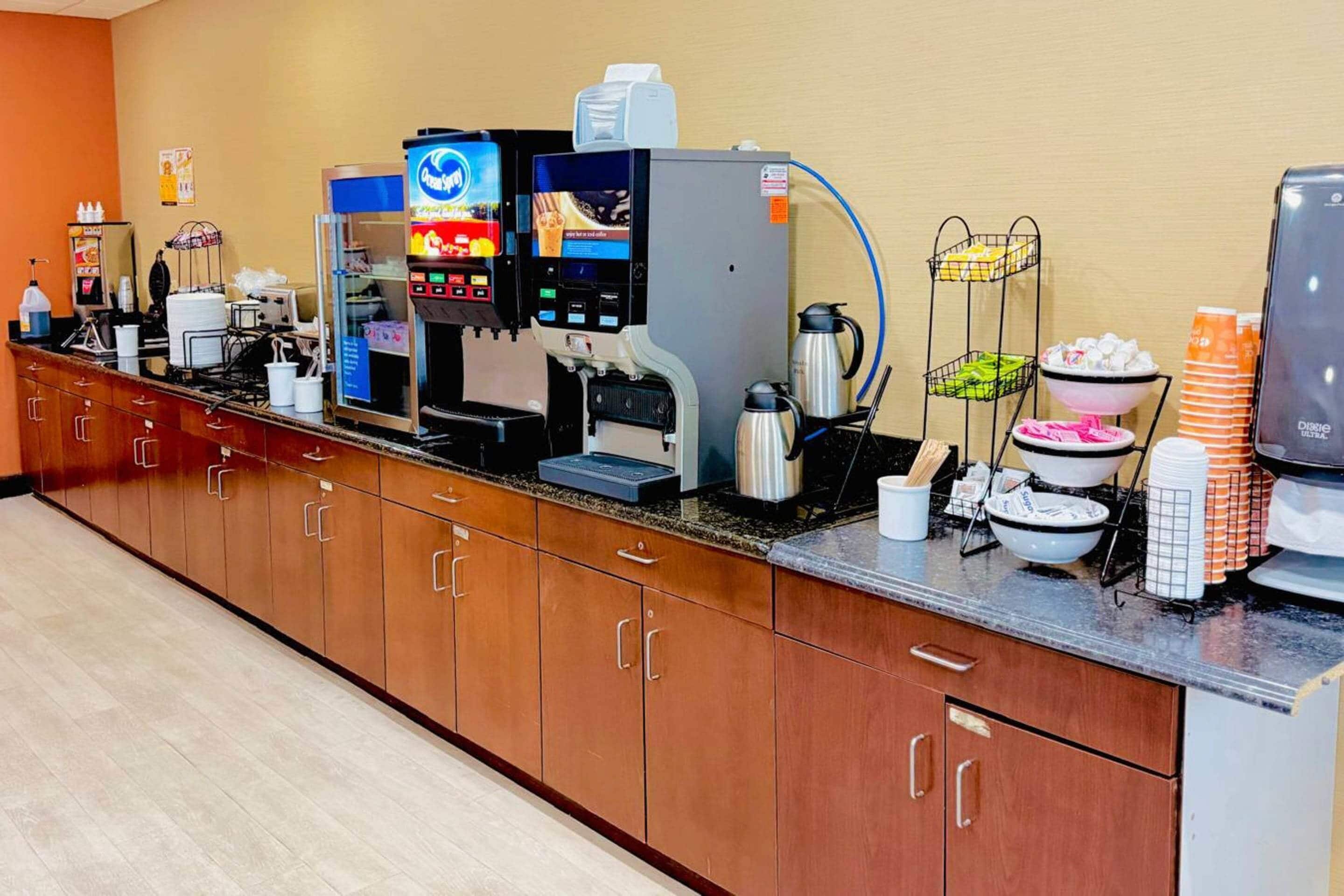 A self-service beverage station with multiple coffee machines, a refrigerated display case, and a variety of disposable cups and containers.
