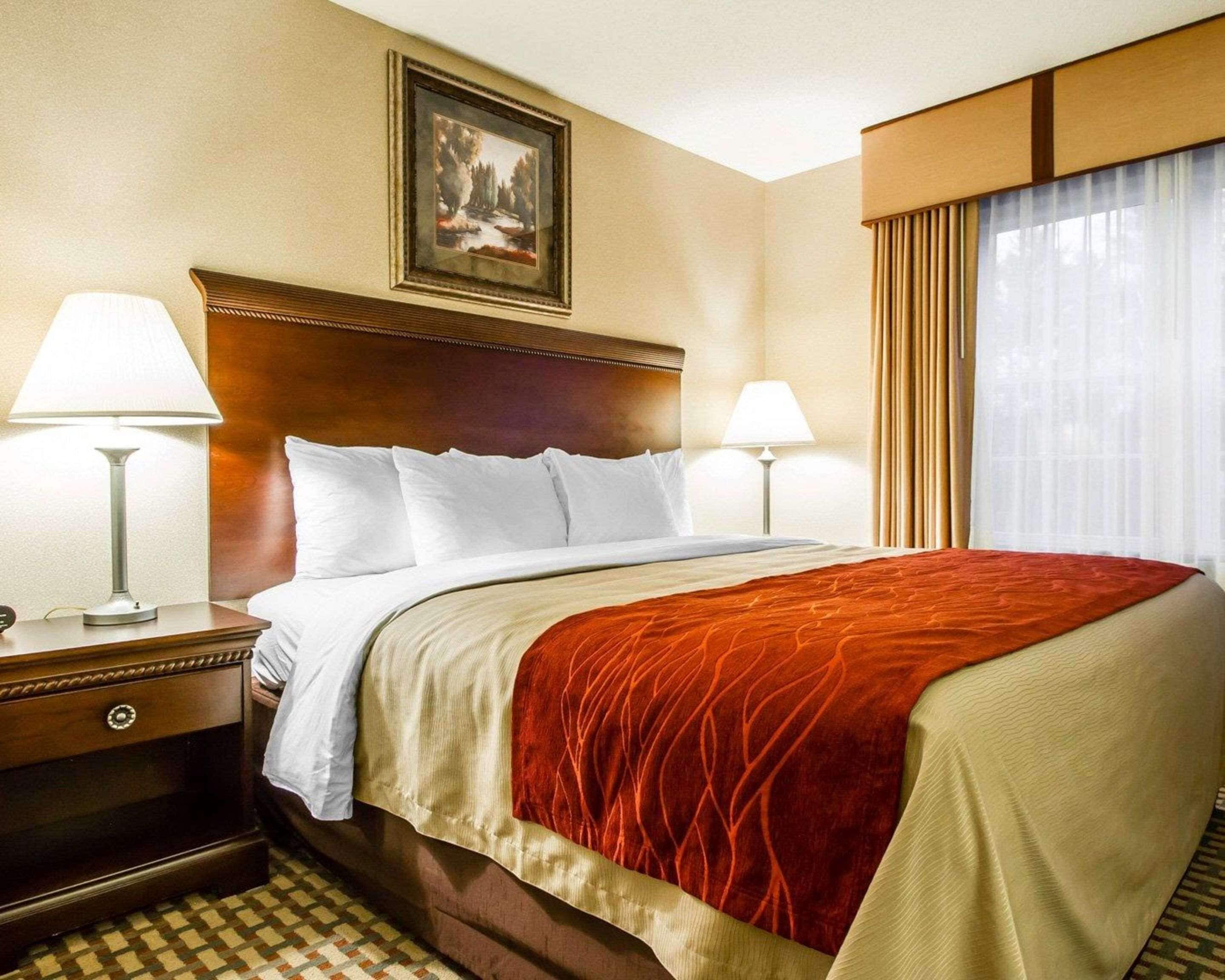 A hotel room with a large bed, two bedside lamps, a wooden headboard, a framed picture on the wall, and a window with curtains.