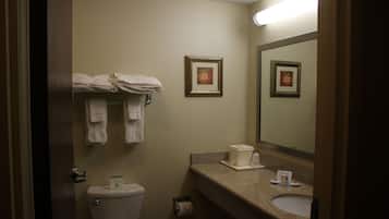 Un baño con inodoro, lavamanos y espejo. Hay toallas colgadas en un perchero y un cuadro en la pared.
