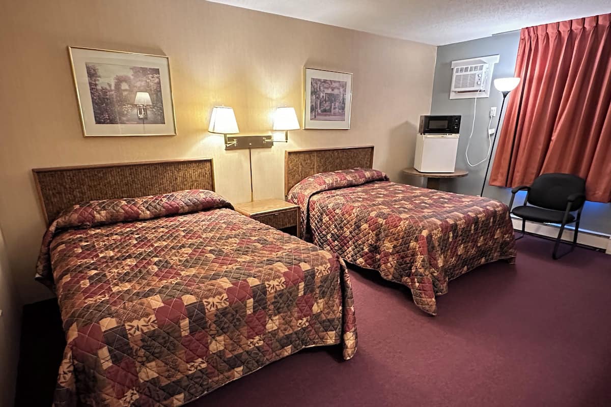 A hotel room with two beds, a nightstand, a lamp, a chair, and a wall-mounted air conditioner.