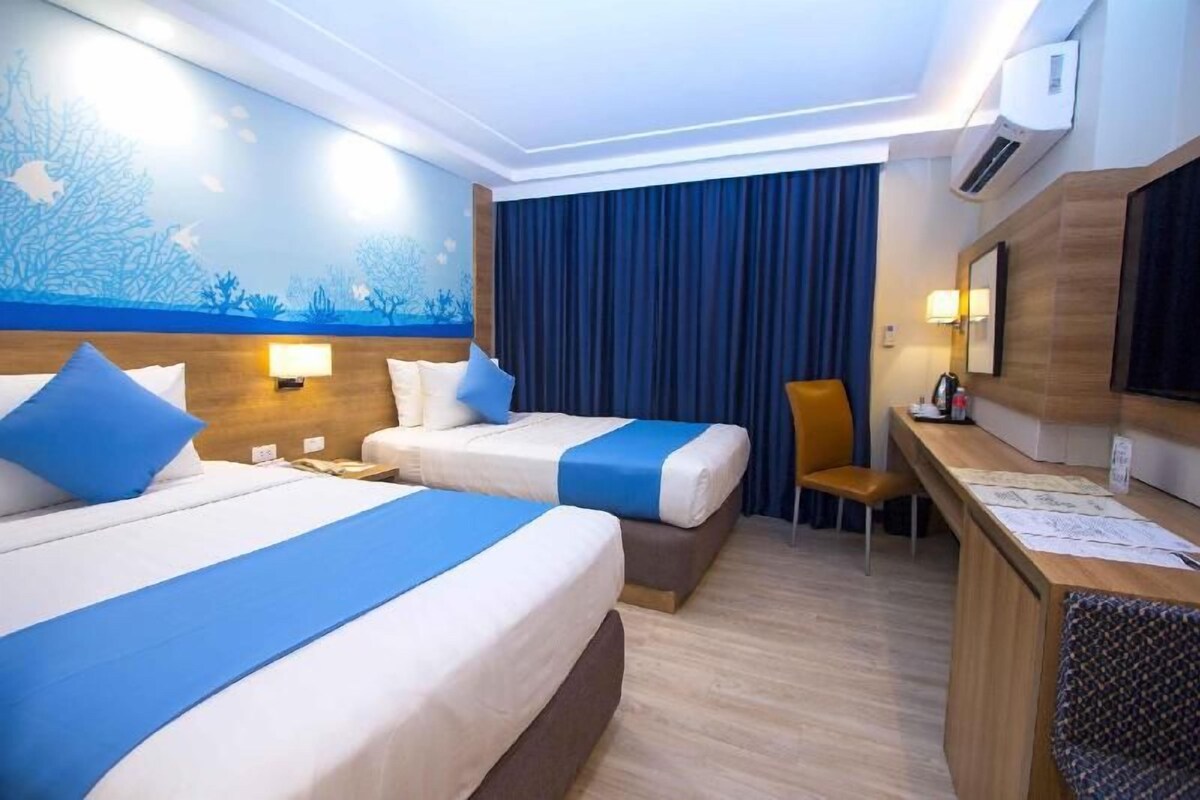 A hotel room with two beds, a desk, a chair, a large window with curtains, and a wall mural.