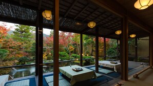 Um quarto tradicional japonês com piso de tatame, uma mesa baixa com toalha e vista para um jardim com lago e folhagem de outono.