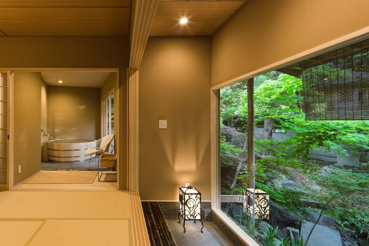 A traditional Japanese room with tatami flooring, a futon, a small table, and a view of a lush garden through a large window.