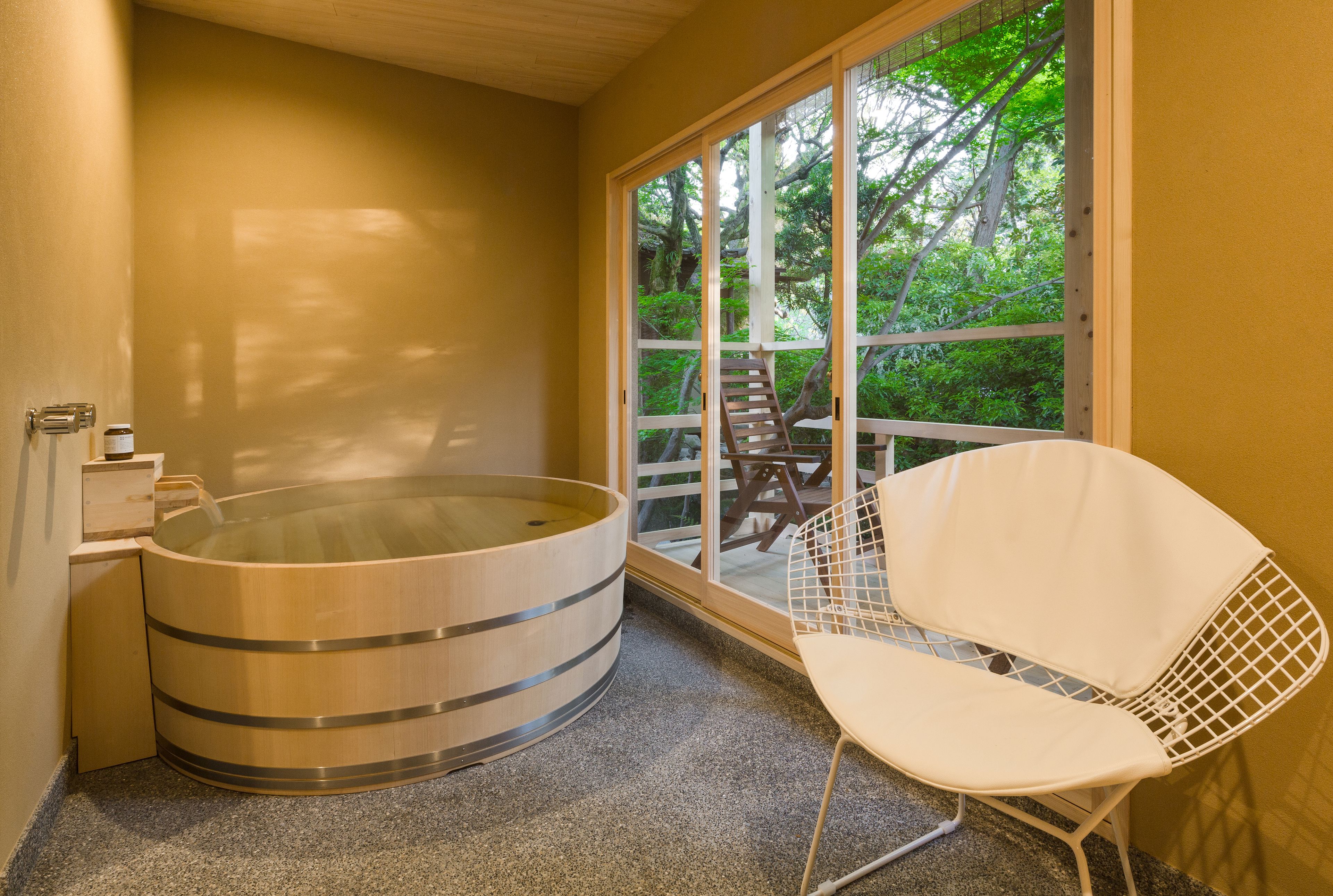 Un cuarto de spa con una bañera de hidromasaje de madera, una silla blanca y un ventanal con vistas a árboles.