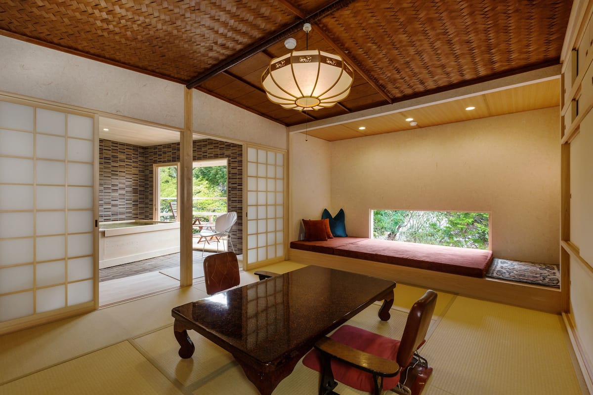 A traditional Japanese room with tatami flooring, a low wooden table, a cushioned bench with pillows, and a sliding door leading to a balcony with a view of greenery.