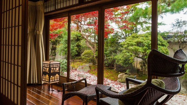 Um quarto japonês tradicional com piso de tatame, um painel shoji, uma mesa baixa com almofada e vista para um jardim com folhagem avermelhada.