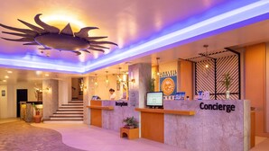 Un espace d’accueil d’hôtel comprenant un comptoir de réception portant l’inscription « Concierge », un autre comptoir de réception portant l’inscription « Réception », et un mur sur lequel est affiché un panneau indiquant « SOLDEIL BORACAY ».