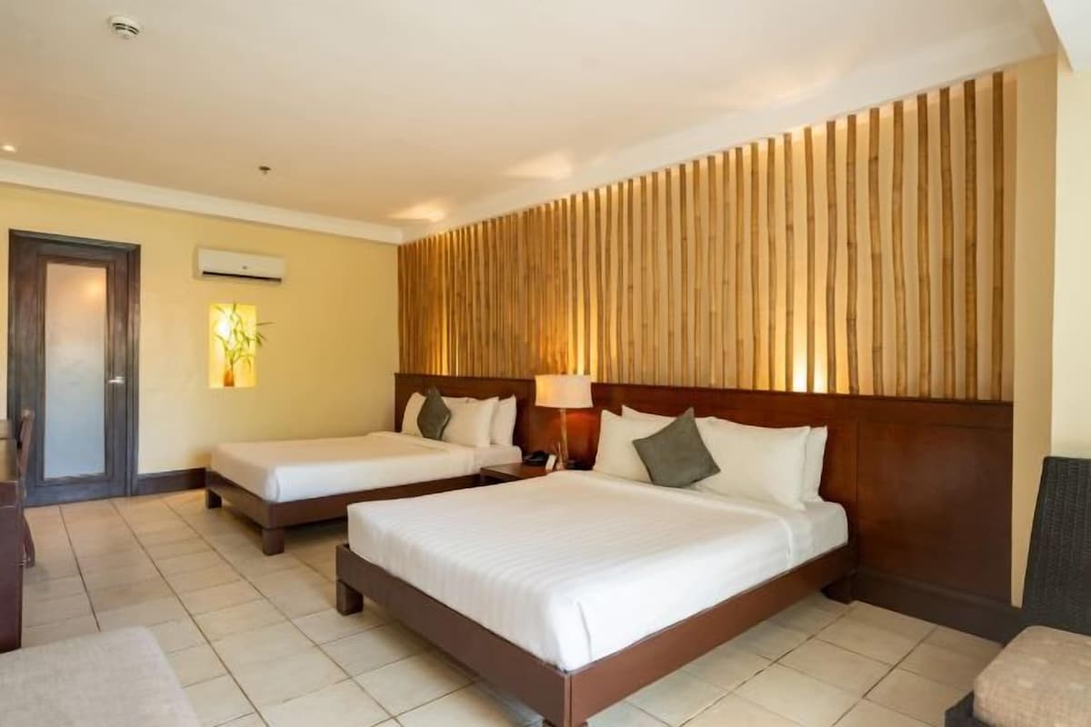 A hotel room with two beds, a wooden headboard, a bedside table with a lamp, and a door.