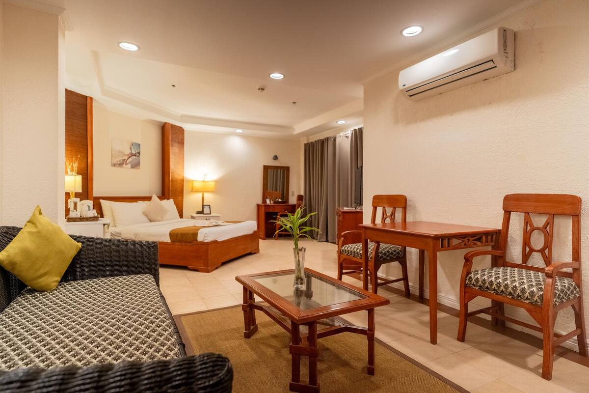 Hotelkamer met een bed, een sofa, een eettafel met stoelen, een bijzettafeltje met een vaas en een airconditioner aan de muur.