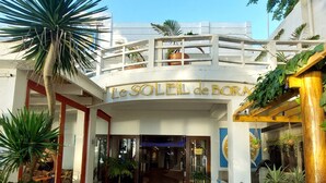L’entrée d’un hôtel, avec une enseigne indiquant « Le Soleil de Bora », une allée couverte et des plantes en pot.