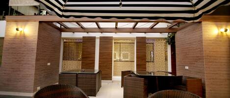 A covered outdoor seating area with wicker furniture and a glass table, surrounded by wooden walls and decorative screens.