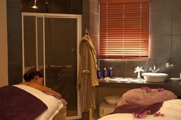 Un cuarto de spa con cabina de ducha de vidrio, tocador con lavamanos y artículos de aseo, y cama con manta púrpura.