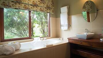 A bathroom with a large bathtub, a wooden vanity, a mirror, and a window with a view of trees.