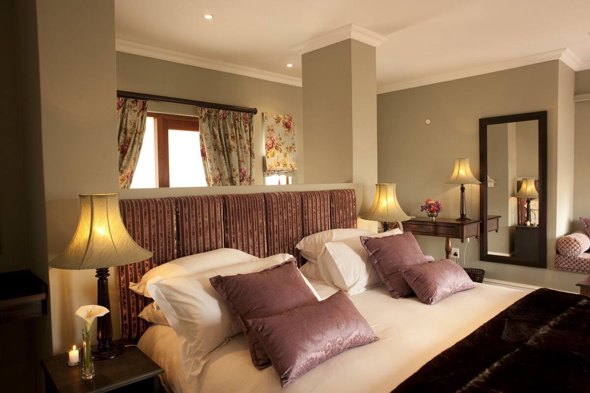 A hotel room with a large bed, two bedside lamps, a nightstand with a candle, a mirror, and a window with curtains.