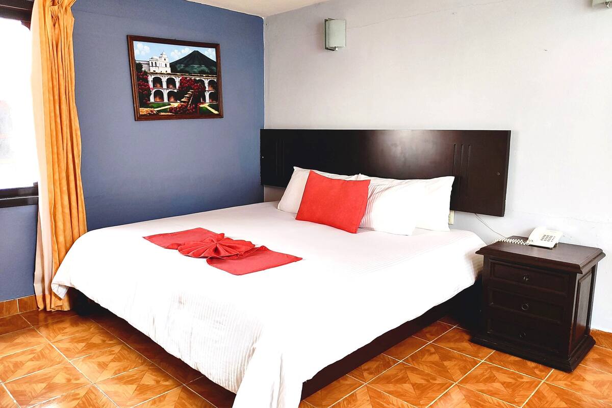Une chambre d’hôtel comprenant un lit, une table de chevet, un téléphone, une lampe fixée au mur et un tableau représentant un bâtiment.