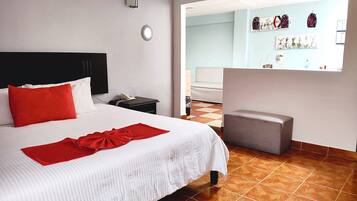 Quarto de hotel com uma cama, mesa de cabeceira com telefone, travesseiro vermelho, cadeira pequena e luminária fixada na parede.