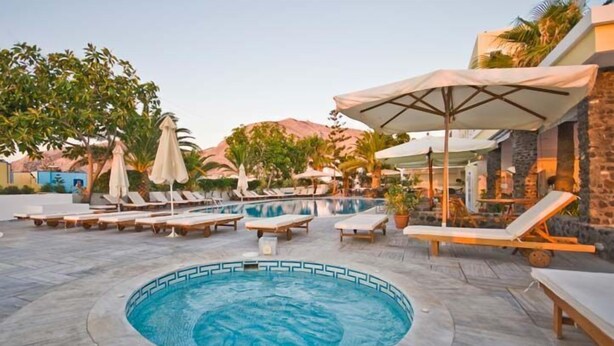 A pool area with lounge chairs and umbrellas, surrounded by trees and a mountain backdrop. - Hotel Makarios (Santorini)