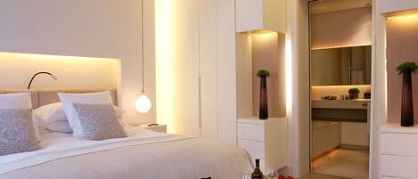 A modern hotel room with a large bed, bedside table, and a nightstand. There is a mirror above the bed, a small plant on the nightstand, and a door leading to a bathroom.