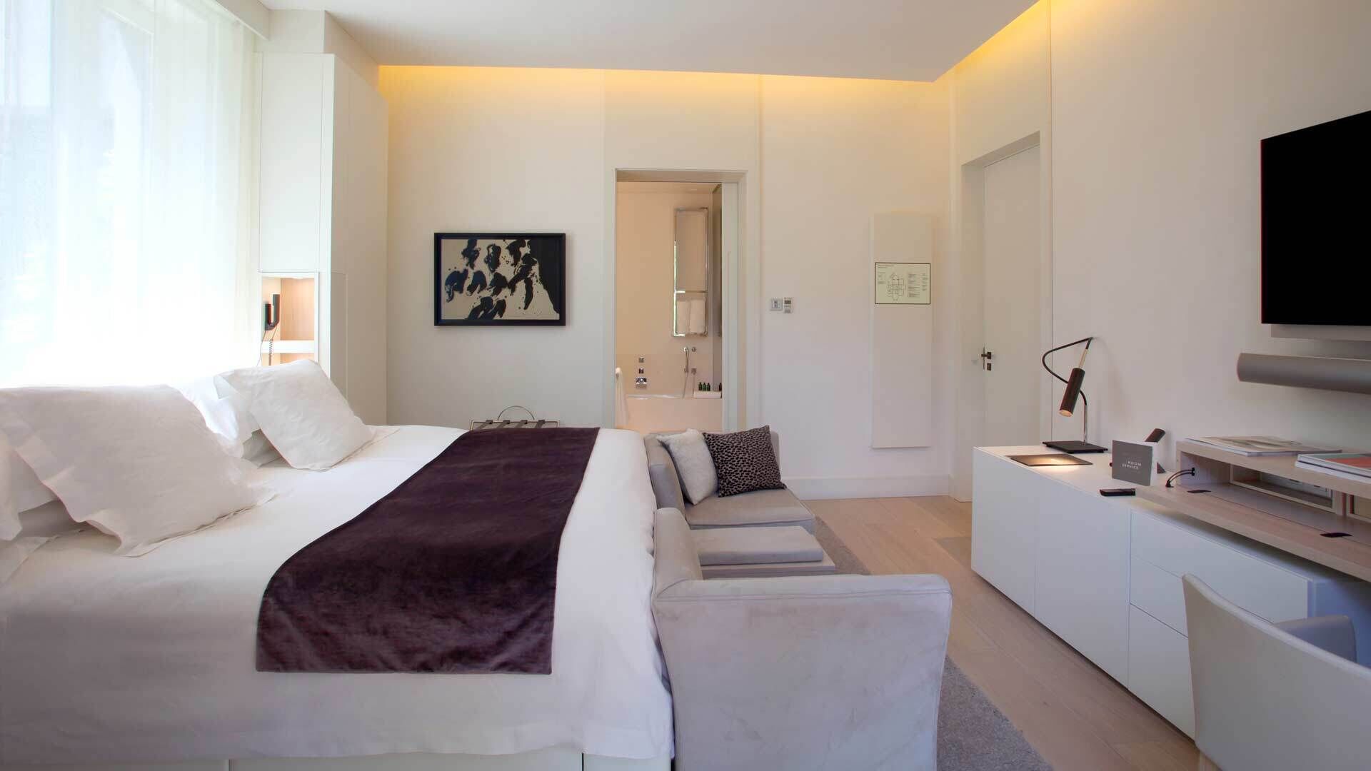 A modern hotel room with a bed, a desk, a television, and a bathroom visible through an open door.