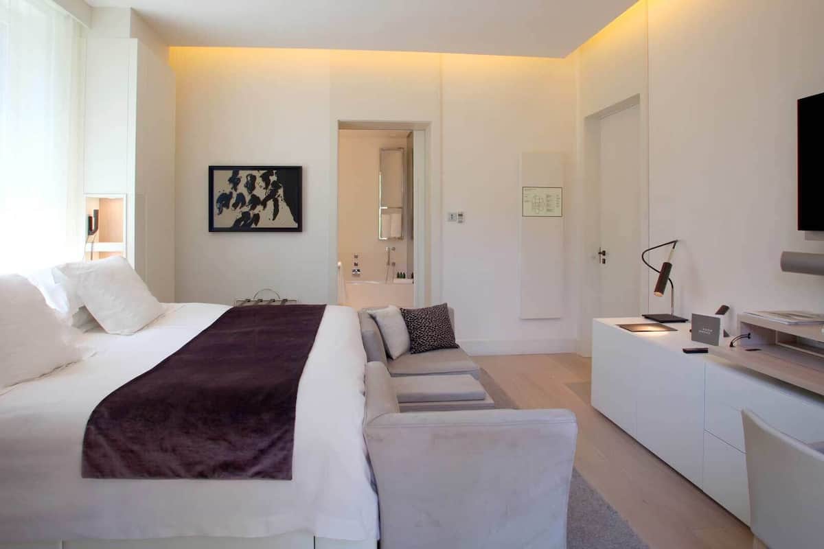 A modern hotel room with a bed, a desk, a television, and a bathroom visible through an open door.