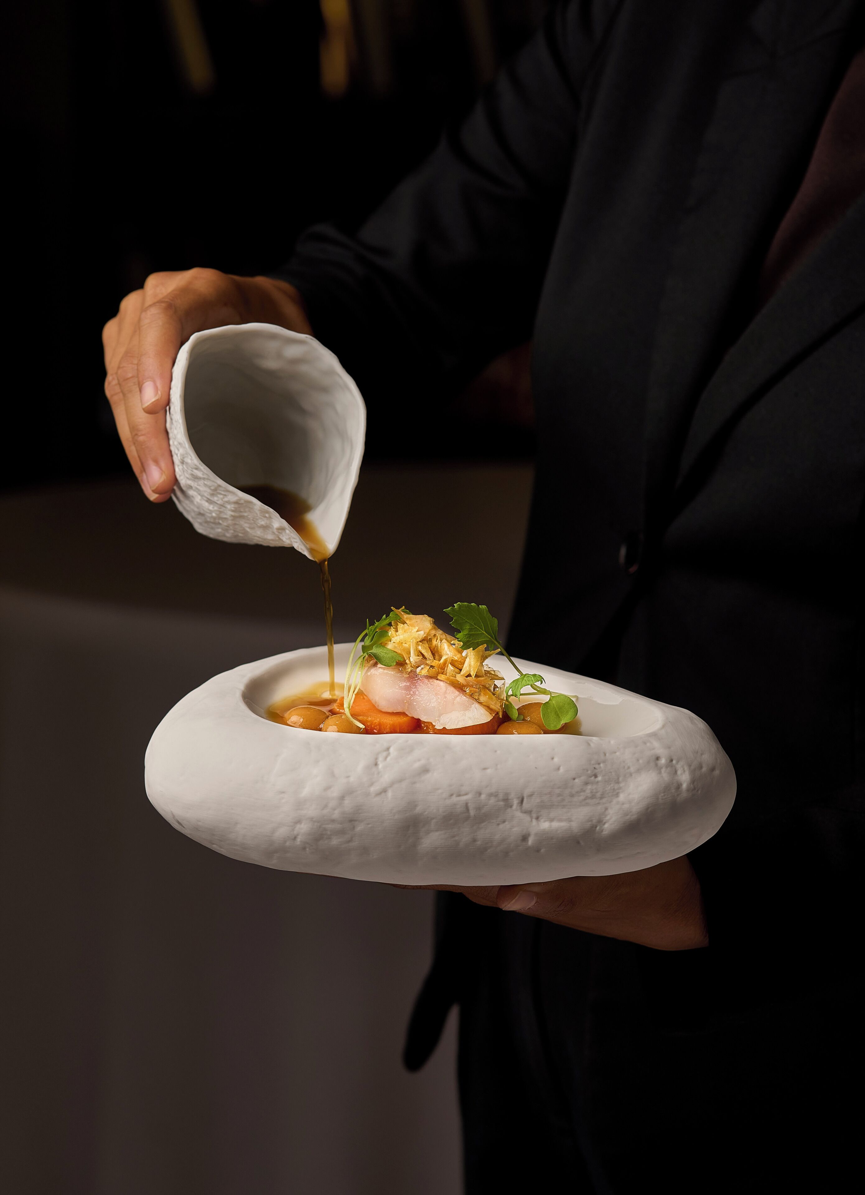 Una persona sosté un plat ceràmic blanc amb un plat a dins, abocant salsa sobre el menjar.