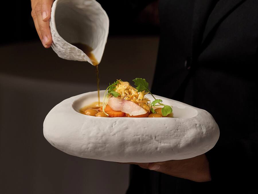 Una persona sosté un plat ceràmic blanc amb un plat a dins, abocant salsa sobre el menjar.