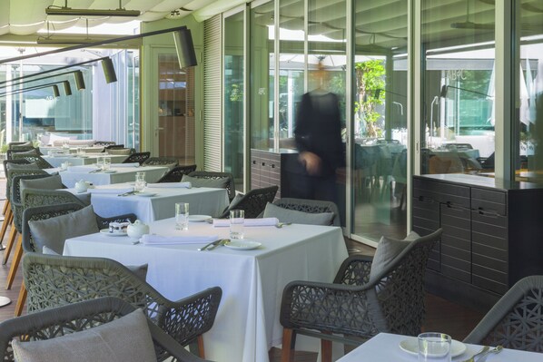 Un área de comedor al aire libre con mesas preparadas para dos, sillas de mimbre y vista a la calle a través de amplios ventanales.