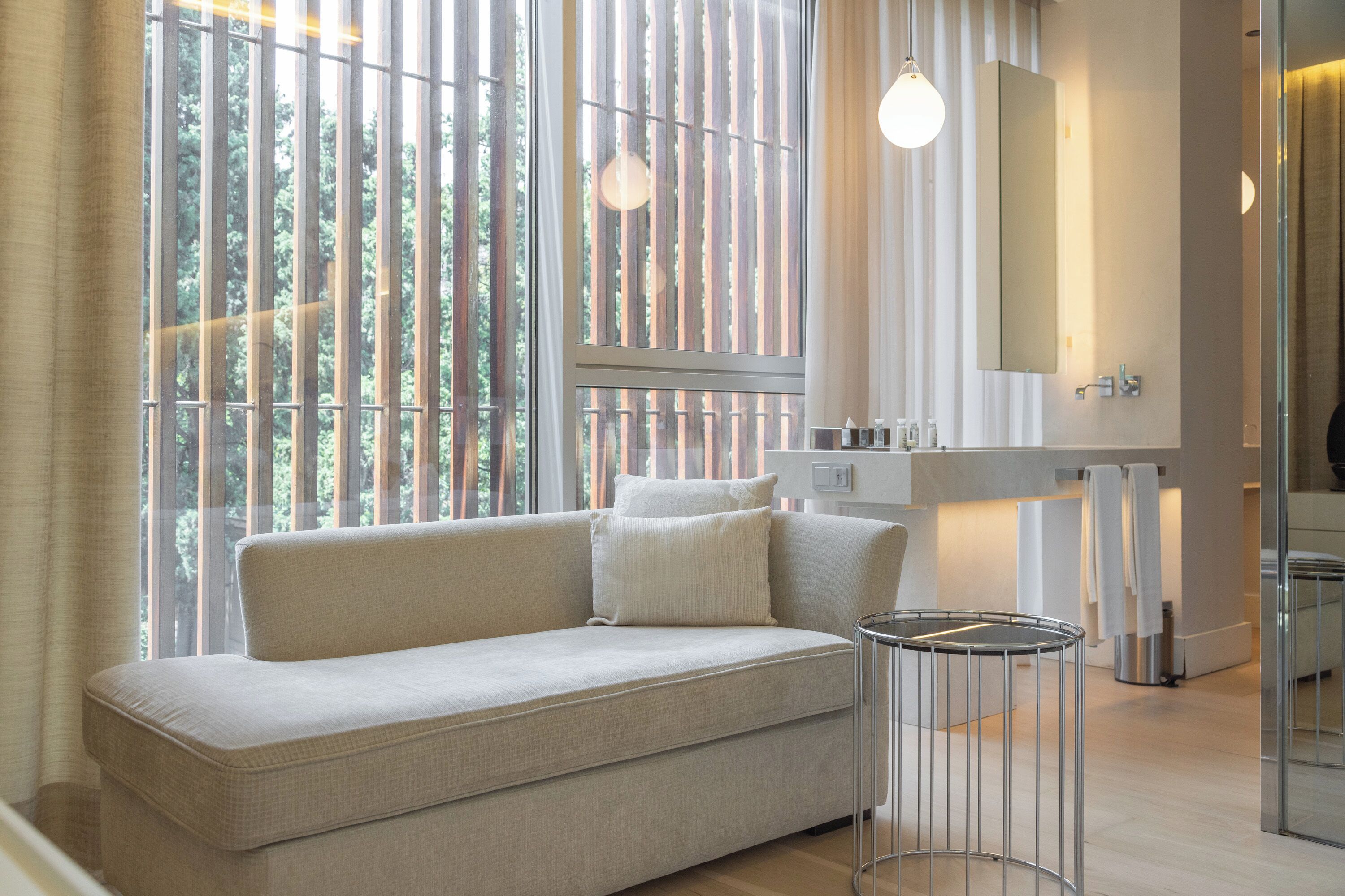Uma sala de estar moderna com um sofá, uma mesa lateral redonda e uma janela grande com persianas verticais.