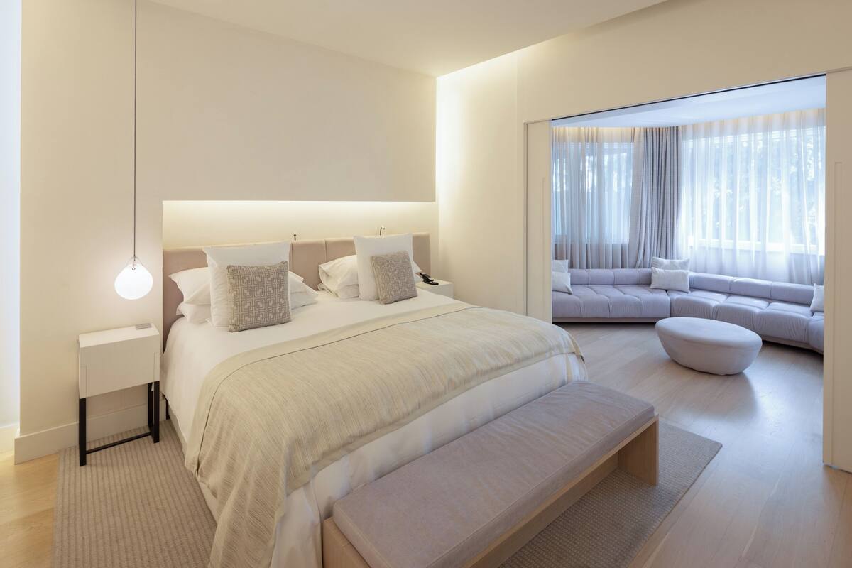 A modern bedroom with a large bed, bedside tables, a bench, and a sofa area with a view.