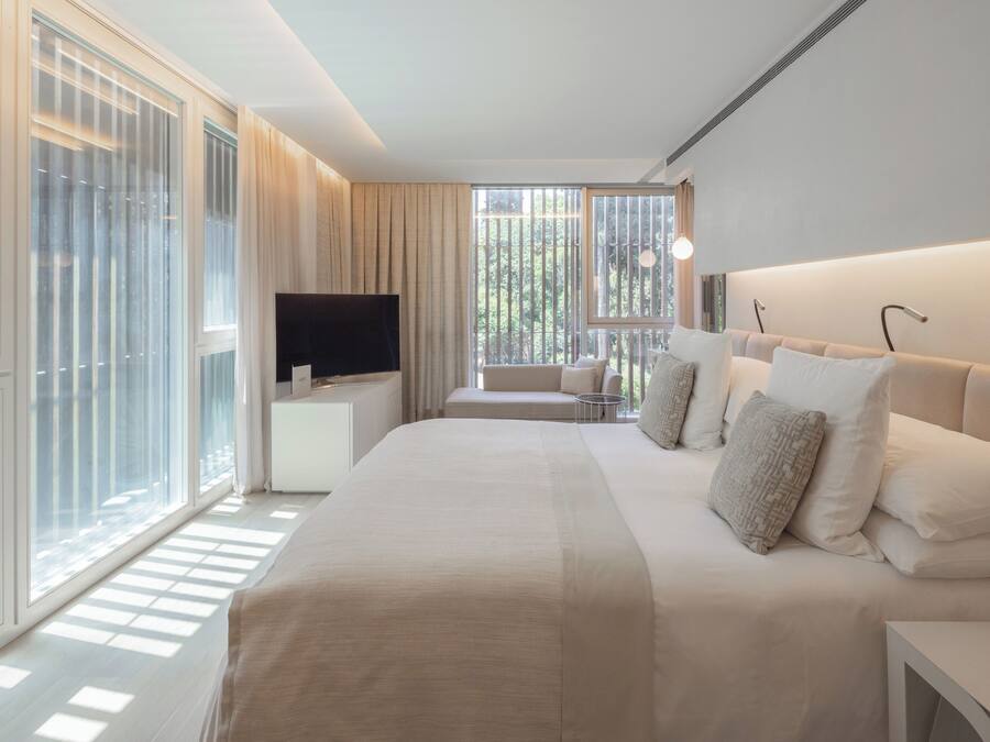 A modern hotel room with a large bed, a TV, and a view of trees through the windows.