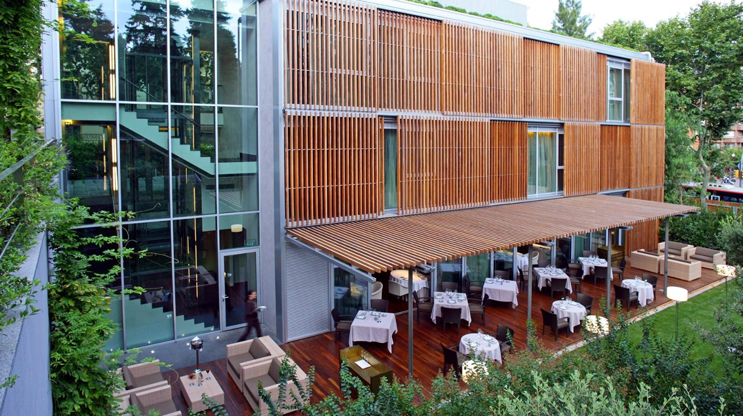 A modern building with a glass facade, wooden slats, and outdoor dining area with tables and chairs.