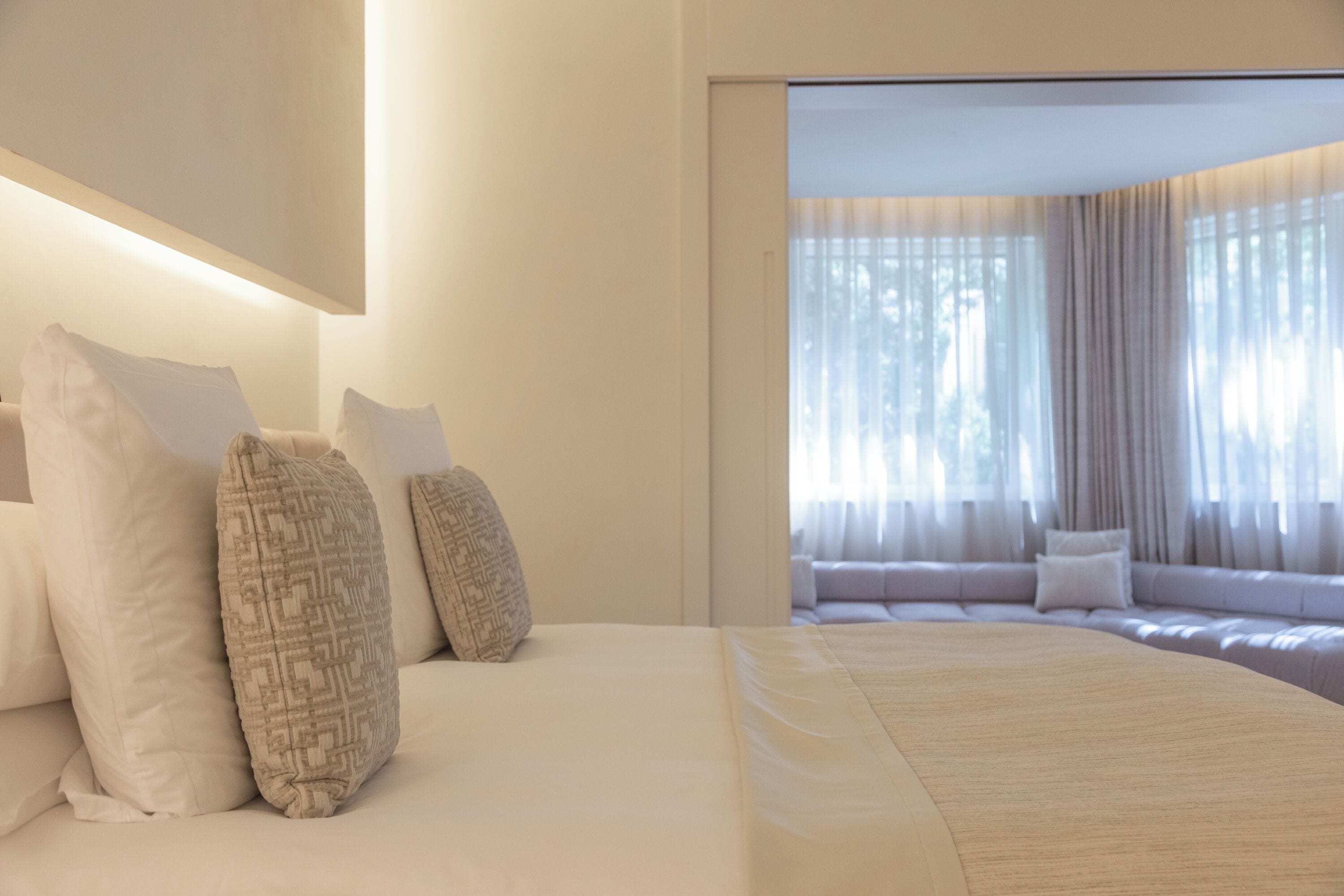 Ein Hotelzimmer mit einem großen Bett, vielen Kissen, einer Couch und Blick durch ein Fenster mit durchsichtigen Vorhängen.