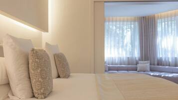 Ein Hotelzimmer mit einem großen Bett, vielen Kissen, einer Couch und Blick durch ein Fenster mit durchsichtigen Vorhängen.