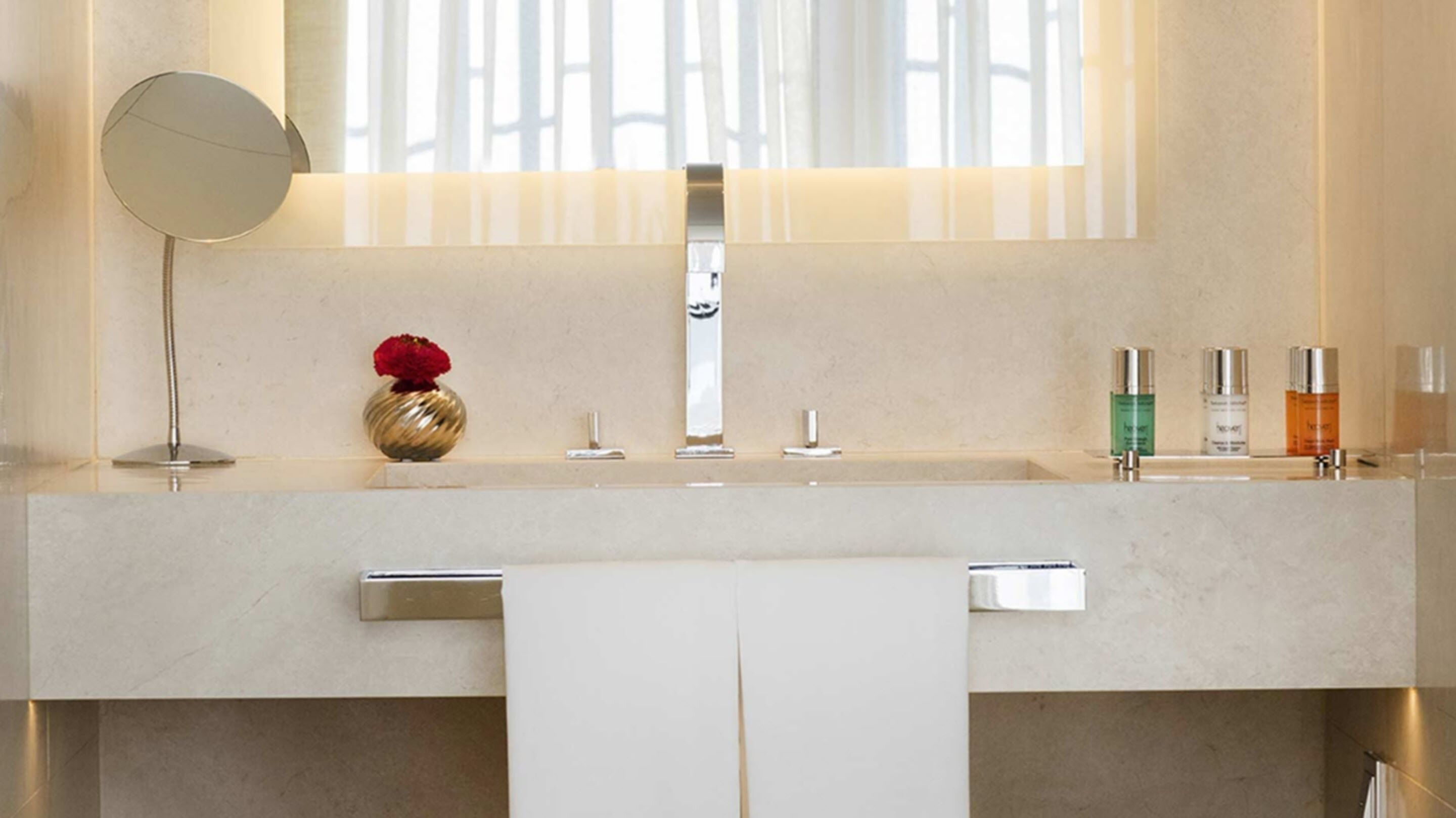 A modern bathroom with a large mirror, a vase with red flowers, and three bottles of toiletries on the sink.