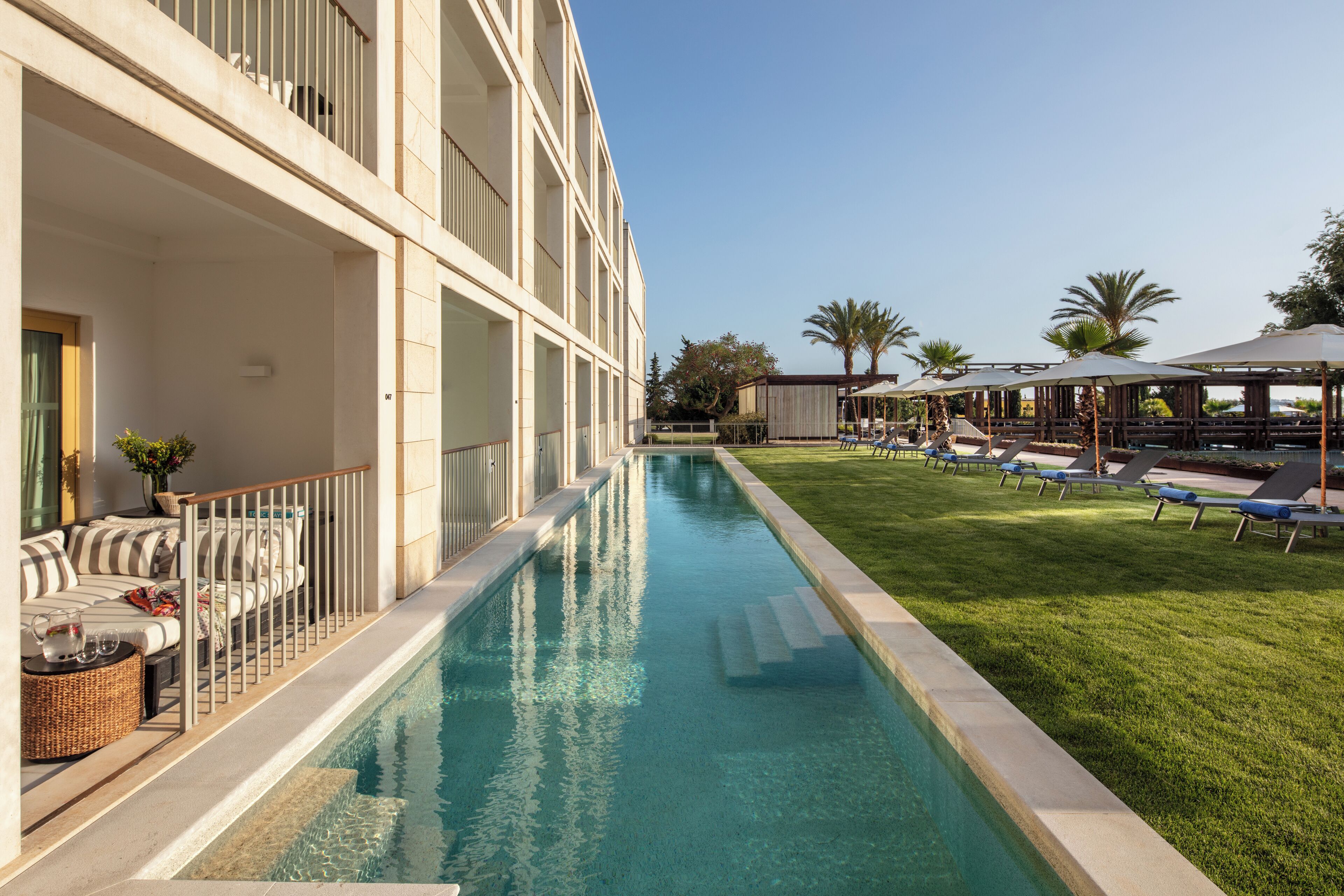 A swimming pool adjacent to a building with balconies, a grassy area with sun loungers, and a covered seating area.